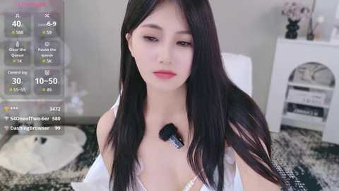 Media: Video of an Asian woman with long black hair, fair skin, and pink lipstick, wearing a white lace bra, holding a black makeup brush. Background shows a modern bedroom with white furniture, a digital screen overlaying stats.