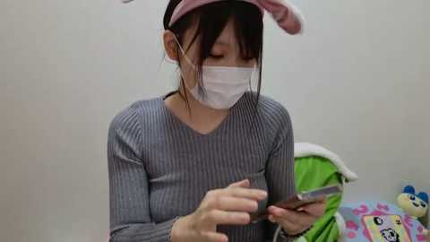 Video of an East Asian woman wearing a gray striped sweater, white mask, and pink bunny hat, examining a phone, in a plain white room with a stuffed animal and a green blanket in the background.