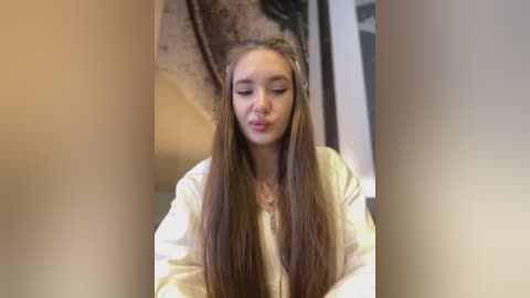 Video of a young Caucasian woman with long, straight brown hair, wearing a white robe, blowing a kiss, in a blurred, beige-toned indoor setting.