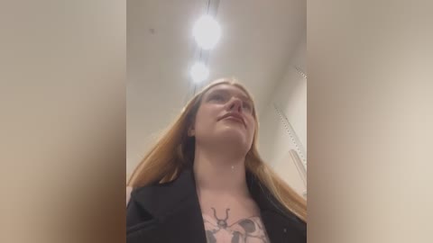 Media: Video of a blonde woman with fair skin, wearing a black blazer, standing in a dimly lit room with a deer tattoo on her chest.