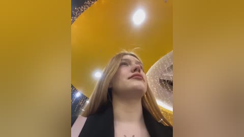 Video of a young woman with long blonde hair, wearing a black jacket, gazing upwards at a cluster of golden and silver balloons in a festive indoor setting, lit by warm overhead lights.