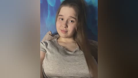 Media: Video of a young Caucasian woman with light skin, straight brown hair, and a gray shirt. She has a gentle smile and is leaning against a blue and black textured wall.