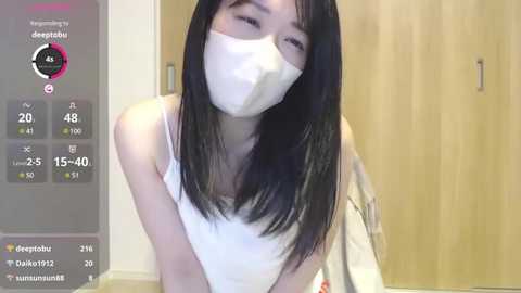 Video of an East Asian woman with straight black hair, wearing a white mask, tank top, and sitting indoors with wooden furniture.