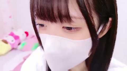 Media: Video of an East Asian woman with straight, dark hair and blunt bangs, wearing a white face mask, looking downward, in a soft, pastel-colored room with a plush Hello Kitty toy.