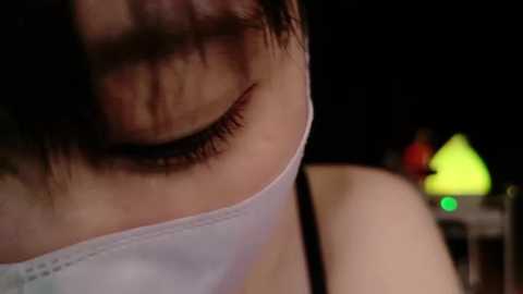 A close-up video of a person with short, dark hair wearing a white surgical mask, highlighting their eyes and nose, in a dimly lit indoor setting.