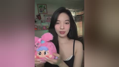 Media: A video of an Asian woman with long black hair and fair skin, wearing a black spaghetti-strap top, holding a pink plush toy with blue hair. The background features a cluttered room with framed pictures and a metal shelf.