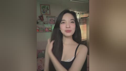 Media: Video of an Asian woman with long black hair, wearing a black camisole, posing indoors with framed artwork and stuffed toys in the background.