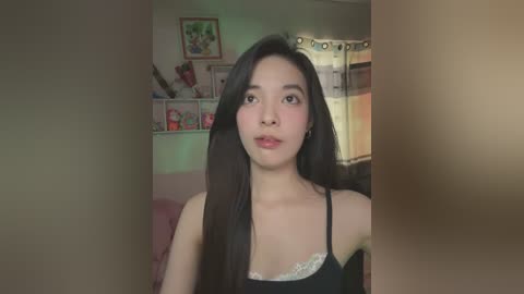 Media: Video of a young Asian woman with long black hair, wearing a black spaghetti-strap top, in a cozy, dimly-lit bedroom with pastel-colored walls and a decorative shelf.