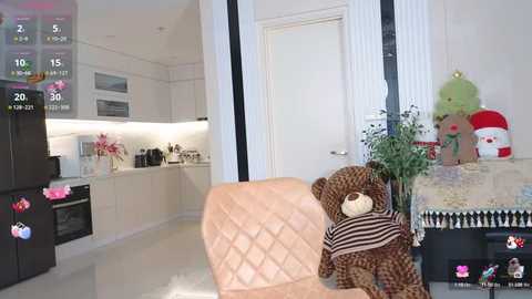 Media: Video of a modern kitchen with a plush brown bear wearing a striped shirt, positioned in front of a black refrigerator. The background features white cabinets, a door, and a decorative Christmas tree.