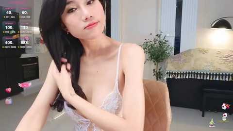 Media: Video of a slender, light-skinned Asian woman with long black hair, wearing a sheer white lace bra, sitting in a modern living room with a beige couch and a green plant.