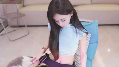 Media: Video of an East Asian woman with long black hair, wearing a light blue crop top, sitting on a chair, playing with a grey and white cat.