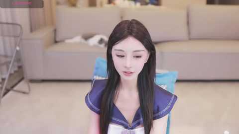 Media: Video of a young East Asian woman with pale skin, long black hair, and delicate features, wearing a sailor school uniform, sitting on a blue stool in a modern living room with beige furniture and a cluttered background.