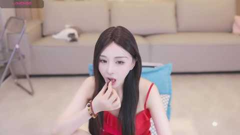 Media: Video of a young East Asian woman with long black hair, fair skin, and slender figure, wearing a red strapless dress, licking a finger while seated on a blue chair in a modern living room.