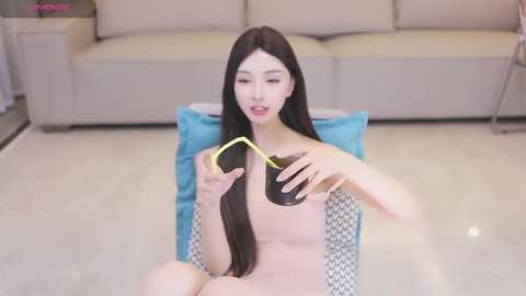 Media: Video of an East Asian woman with long black hair, fair skin, and slender physique, sitting on a light blue chair, sipping tea from a black cup with a yellow straw, in a modern living room with beige sofa and blue pillows.