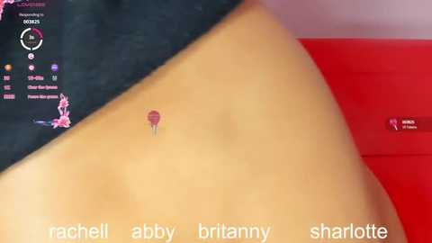 A video of a woman's tan, smooth thigh with a small, red balloon piercing on her inner thigh. She wears black shorts. The background includes a red sofa and a pink wall.