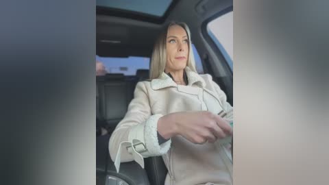 Media: Video of a blonde woman in a beige coat, driving a car. The car interior is visible, showing a steering wheel and dashboard.