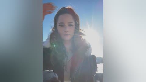 Media: A video of a young woman with wavy brown hair and a fur-lined coat, standing in front of a blurred, bright, possibly snowy landscape, with a sunburst effect in the background.