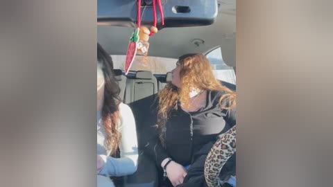 Media: Video of a woman with long curly red hair, wearing a black hoodie, sitting in a car, looking out the window, with a leopard-print scarf draped over her shoulder.