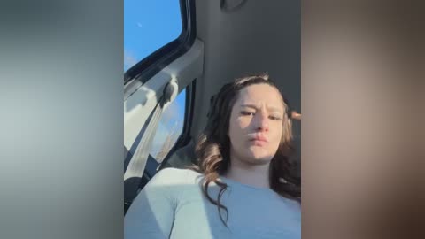 Media: Video of a woman with long, wavy brown hair, wearing a white shirt, seated in a car with a blue sky visible through the window.