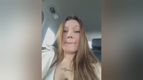 Media: A video of a young Caucasian woman with long, straight, light brown hair, wearing a white hoodie, seated in a car. She has a tattoo on her chest. The background shows the car's interior.