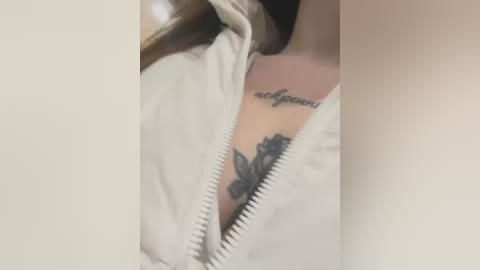 Video of a partially unzipped white jacket revealing a tattoo of a black rose with a heart-shaped center on a person's chest, with light skin and shoulder-length brown hair visible.