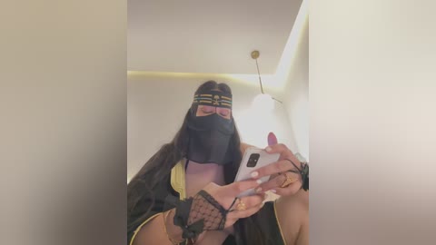 Media: Video of a woman with long dark hair, wearing a black mask, black outfit with gold accents, and a headband, holding a smartphone, in a modern, dimly lit room with a ceiling light.