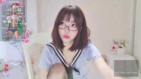 Media: Video of a young Asian woman with straight black hair, glasses, and a blue sailor school uniform, sitting in a room with a white table and floral decorations.