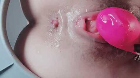 A close-up video shows a woman's vulva with a pink, glossy silicone toy inserted into her anus. Her skin is light, and there is visible moisture. The background is out of focus.