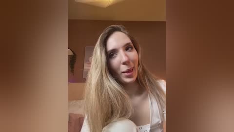 Media: A video of a young, smiling Caucasian woman with long, straight blonde hair, wearing a white blouse, in a warmly lit room with beige walls.