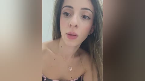 Media: Video of a young Caucasian woman with long, straight brown hair, light skin, and subtle makeup, wearing a strapless, patterned top. Her expression is neutral, and the background is blurred, focusing on her face.