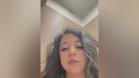 Media: A close-up video of a woman with medium brown skin and curly hair, wearing minimal makeup, captured from a low angle. The background features beige-tiled walls and a ceiling.