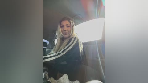 Media: Video of a woman with long, straight hair in a black jacket and white scarf, sitting in a car, looking down. Bright lens flare, dim interior light.