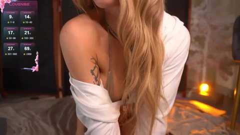 Media: Video of a blonde woman with long wavy hair, wearing a white off-shoulder top, revealing a tattoo on her left shoulder. Background shows a dimly lit room with a fireplace, a TV displaying a weather forecast, and a cozy ambiance.