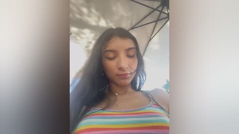 Media: Video of a young woman with long dark hair, wearing a colorful striped tank top, standing under an umbrella in bright daylight.