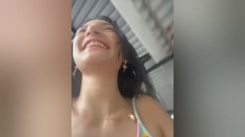 Video of a smiling young woman with medium skin tone and dark hair, wearing a colorful tank top, in a modern indoor setting with industrial ceiling.