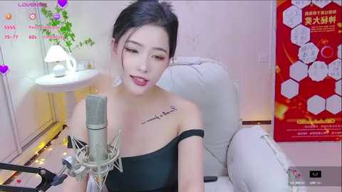 Media: Video of an East Asian woman with dark hair, wearing a black off-shoulder top, singing into a microphone in a cozy room with a white chair, green plant, and a red calendar.