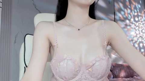 Media: Video of a fair-skinned Asian woman with straight black hair, wearing a sheer pink lace bra with thin straps, sitting on a white chair against a marble wall with a chandelier.