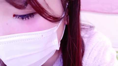 Video of a person with long, dark brown hair wearing a white surgical mask, showing close-up of their eye with visible redness and slight swelling, suggesting possible irritation or infection.