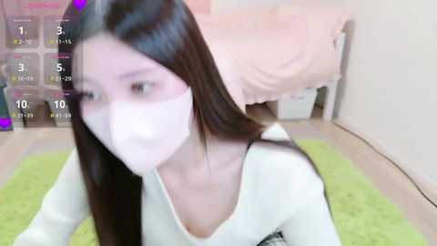 Media: A blurry video of a young Asian woman with long black hair and a white face mask, wearing a white top, sitting on a green rug in a pink bedroom with a bed.