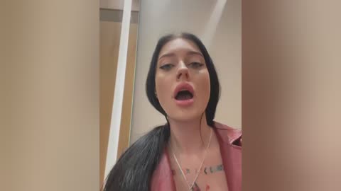 Video of a young woman with long black hair, wearing a pink jacket, in a beige bathroom stall, mouth open, looking distressed.