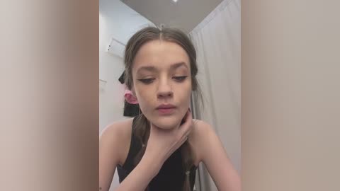 A video of a young girl with long, light brown hair in pigtails, wearing a black tank top, looking down at her hands in a narrow, white-walled bathroom with a shower curtain in the background.