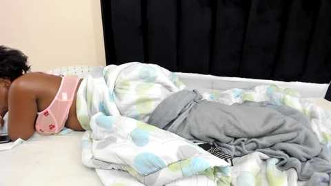 Media: Video of a woman with dark skin, short black hair, lying on a white bed with light blue and green polka dot sheets, wearing a pink tank top and gray pants, under a gray blanket.