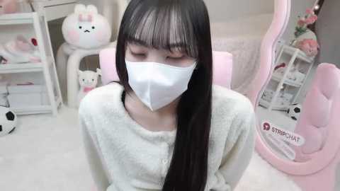 Video of a young Asian woman with long black hair, wearing a white mask, cozy white sweater, and pink chair in a pastel-themed room with stuffed animals and a pink desk.