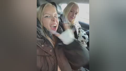 Media: Video of two blonde women in a car; one wearing a brown puffer jacket, the other in a cow-print sweater, both smiling and making a peace sign.