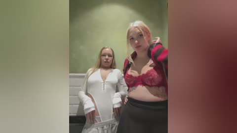 Media: Video of two young women in a bathroom: one in a white zip-up, the other in a red lace bra and black pants. Background shows green walls and white tiles.