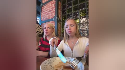 Media: Video of two young blonde women, one in a red-striped shirt, the other in a white zip-up, sitting at a table in a cozy cafe with brick walls and glass block windows.