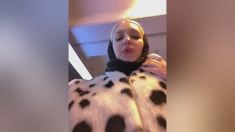 Video of a young blonde woman in a cow costume, wearing a black hood with white spots. She's indoors with fluorescent lighting and a blurred background.