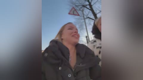 Video of a blonde woman laughing in a grey coat, outdoors with a clear blue sky and a triangular road sign in the background.