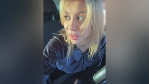 Video of a blonde woman with fair skin and blue eyes, wearing a black shirt, sitting in a car, looking concerned, blurry background.