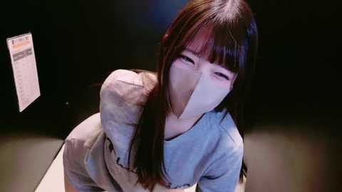 Media: A video of a young Asian woman with long, dark hair, wearing a light grey hoodie and a face mask, crouched down, looking up, in a dimly lit room.